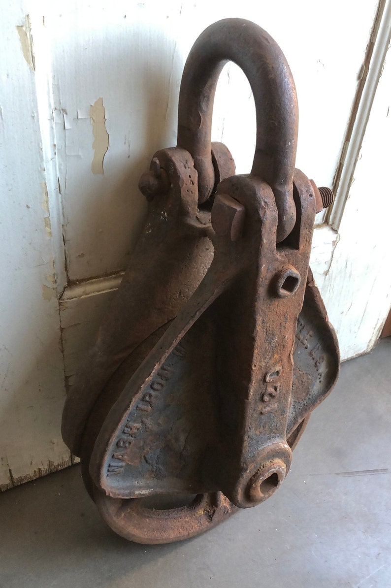 May include: A rusty metal pulley with a large, round wheel and a hook on top. The pulley is labeled "WABASH IRON WKS" and "U.S.A."