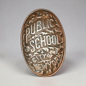 May include: Oval-shaped, antique metal badge with the words "PUBLIC SCHOOL CITY NEW YORK" in raised lettering. The badge has an aged, bronze-like appearance with ornate scrollwork around the text, suggesting a vintage or historical item.