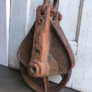 May include: A rusty metal pulley with a square hole in the center and a hook on top. The pulley has the text "MACHINERY" and "220" on the side.