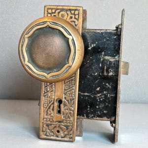 May include: Antique brass door knob and lock set. The knob is round with a decorative edge and a dark center. The lock plate and surrounding hardware feature intricate embossed patterns. The metal has a weathered appearance.