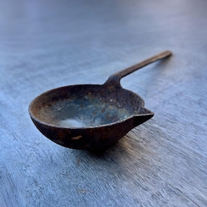 May include: A small, antique metal ladle with a long handle. The ladle has a rounded bowl and a spout. The metal is rusty brown with hints of blue and green. The ladle is likely used for melting or pouring.