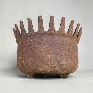 May include: A weathered, rusty brown metal planter with a crown-like rim and three small feet. The planter has a rounded body and a textured surface, suggesting an antique or vintage aesthetic. Ideal for small plants or decorative use.