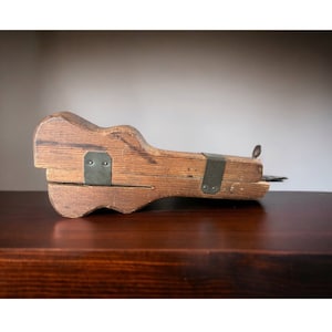 May include: A wooden tool with metal accents, resembling a guitar shape. The tool is made of light brown wood and has two metal bands with rivets. The tool is sitting on a dark brown wooden surface.