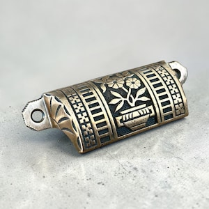 May include: A decorative brass drawer pull with a floral design. The rectangular pull features a detailed pattern of flowers in a pot, surrounded by vertical and horizontal lines. The pull has two mounting holes and a slightly curved shape.