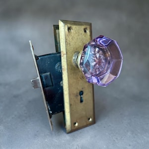 May include: Antique door hardware set featuring a faceted, lavender glass doorknob. The knob is mounted on a brass plate with a keyhole and a dark metal lock mechanism. The hardware shows signs of age and wear.