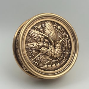 May include: A gold-colored, circular decorative object with a detailed relief of a bird in flight, surrounded by foliage. The object has a raised border and a slightly domed surface, suggesting it could be a lid or a decorative element.