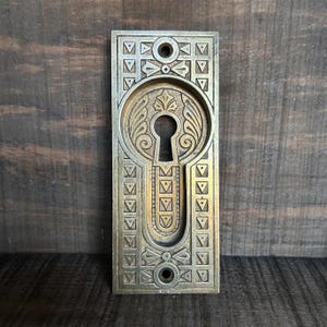 May include: Ornate brass-toned keyhole cover with a rectangular shape. The cover features a keyhole in the center, surrounded by decorative floral and geometric patterns. Two holes are at the top and bottom for mounting. The background is a dark wood.