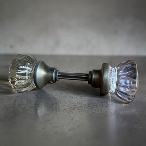 May include: A vintage door knob with clear, faceted glass knobs and a brushed metal finish. The door knob is lying horizontally, showcasing its classic design and potential for home decor or restoration projects.