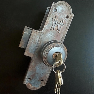 May include: An antique, weathered metal door lock with a key inserted. The lock features an ornate design with the initials "WK" and decorative edging. The keys are on a metal ring. The lock has a distressed, aged appearance.