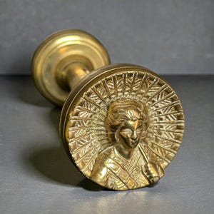May include: A vintage brass door knob with a detailed relief of a figure surrounded by a sunburst design. The knob has a cylindrical handle and a circular face. The brass has a warm, golden hue.
