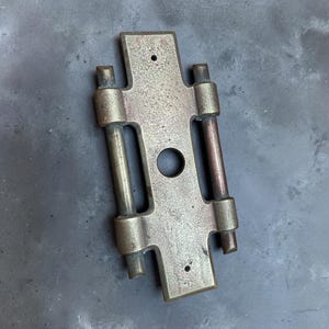 May include: A vintage brass door hinge with a rectangular plate and two cylindrical hinge pins. The metal has a tarnished, aged appearance, with a central circular hole and two smaller holes. The overall design is symmetrical.