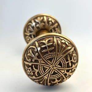 May include: Close-up of two antique brass door knobs. The knobs feature intricate floral and geometric patterns. The brass has a warm, aged patina, suggesting vintage craftsmanship. The design is symmetrical and detailed, showcasing a classic aesthetic.
