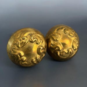 May include: Two antique brass-colored spherical knobs with ornate, swirling relief patterns. The knobs have a warm, golden hue and are likely for furniture or decorative use. The design features detailed, flowing lines.