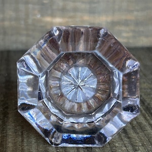 May include: A clear, faceted glass doorknob with an octagonal shape. The center features a starburst design. The doorknob is set against a textured, brown wooden surface, highlighting its reflective qualities.