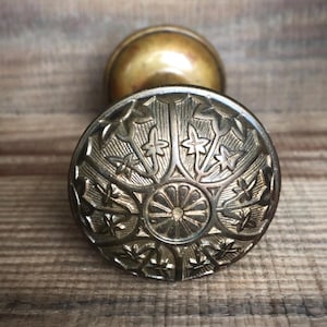May include: Close-up of an antique brass doorknob. The knob features an ornate, embossed design with floral and star-like patterns. The back portion of the knob is a smooth, rounded shape. The background is a wooden surface.