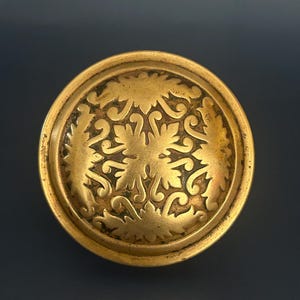 May include: A round, antique-style brass knob with an ornate, symmetrical leaf and scroll design. The knob has a slightly raised edge and a tarnished finish, giving it a vintage appearance. The design is centered on the knob's face.