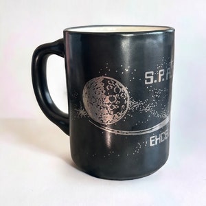 May include: Black ceramic mug with a silver design of a moon and stars. The text "S.P.A.C.E" and "EHCS" is printed on the mug.