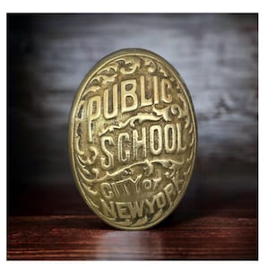 May include: An oval-shaped brass badge featuring raised lettering that reads PUBLIC SCHOOL CITY OF NEW YORK. The metal surface has a weathered, antique finish with decorative scrollwork patterns surrounding the embossed text.