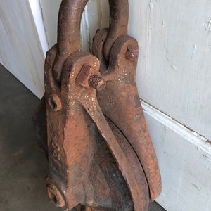 May include: A rusty metal pulley with a large, round wheel and a hook on top. The pulley is made of cast iron and has a rough, textured surface.