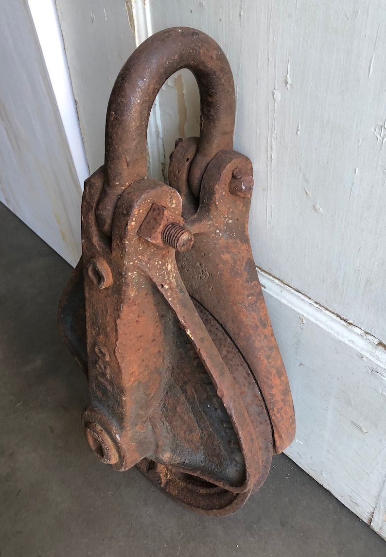 May include: A rusty metal pulley with a large, round wheel and a hook on top. The pulley is made of cast iron and has a rough, textured surface.