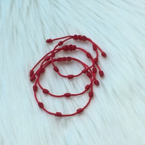 May include: Four red string bracelets with knotted details, arranged in a circle on a white fuzzy surface.