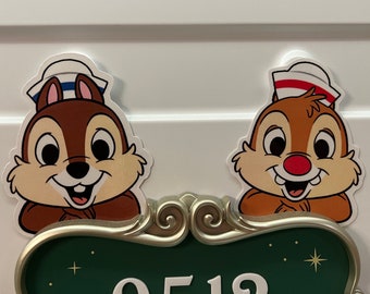 Puede incluir: Dos ardillas de dibujos animados, Chip y Dale, con gorros de marinero, aparecen sobre un letrero verde con el número "2519". El letrero tiene un marco decorativo de color dorado. Las ardillas tienen pelaje marrón y caras sonrientes.