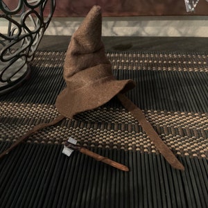 May include: A brown felt wizard hat with a pointed crown and a wide brim. The hat has long, attached ties. A small, wooden wand with a silver tip rests nearby. The items are on a dark, textured surface.