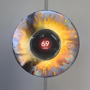 May include: A smart thermostat with a circular design. The display shows the temperature as 69 degrees. The thermostat is set against a decorative metal plate with a sunburst pattern in gold and blue hues.