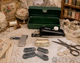 May include: A vintage black Singer handheld sewing machine with a white dial, a green plastic storage case, four grey plastic buttonhole attachments, a metal seam guide, a rectangular white block, and silver metal scissors on a lace cloth.