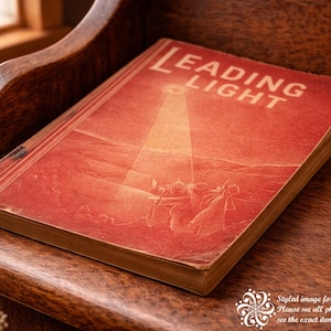 May include: A vintage book titled "LEADING LIGHT" with a red cover and gold-toned edges, resting on a wooden surface. The cover features an illustration of figures under a beam of light. The book is placed on a wooden chair.