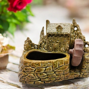 May include: A ceramic planter shaped like a small cottage with a stone wall and a water wheel. The planter is green and brown and has a small opening for planting.