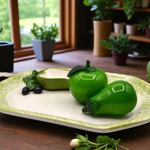 May include: A decorative serving tray with a white center and a light green, speckled border. The tray holds a green apple, a pear-shaped object, a small bowl, and black decorative berries. The objects are glossy and sit on a wooden surface.