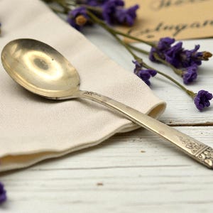May include: A silver spoon with a rounded bowl and a decorative floral design on the handle. The spoon rests on a cream-colored linen napkin, with sprigs of purple flowers and a vintage card in the background.