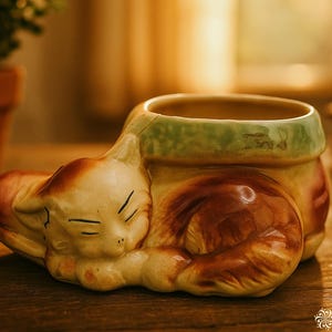 May include: A ceramic cat planter in cream, brown, and green. The sleeping cat is curled around the planter, with its head resting on the side. The planter has a green rim and is sitting on a wooden surface.
