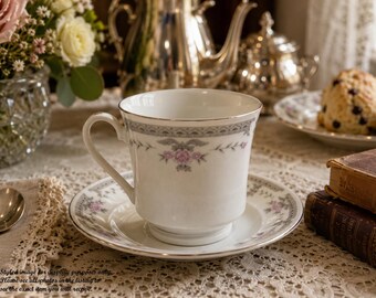 May include: A white porcelain teacup and matching saucer featuring a delicate floral pattern with small pink roses, grey foliage, and a thin silver rim. The teacup has a curved handle and a pedestal base, with the floral design repeated on the saucer.