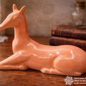 May include: A ceramic deer figurine in a resting pose, with a smooth, glossy finish. The deer is a warm peach color and is lying down on a wooden surface. The figurine has a classic, elegant design.