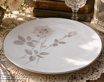 May include: A round white porcelain dinner plate featuring a delicate gray botanical illustration of a blooming rose and a rosebud with leaves. The plate has a thin metallic silver rim around the edge, providing a clean and polished finish.