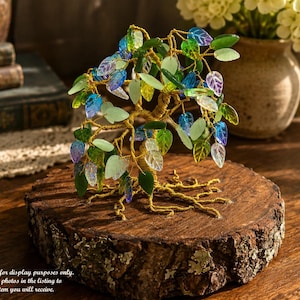 May include: A decorative wire tree sculpture featuring a twisted gold-colored metal trunk and branches adorned with numerous translucent, leaf-shaped glass beads in shades of blue, green, and purple, mounted on a circular natural wood slice base.