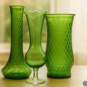 May include: Three green glass vases of varying shapes and sizes. The vases have textured diamond patterns and a smooth, scalloped edge. The vases are displayed on a wooden surface with a blurred background of a window and greenery.
