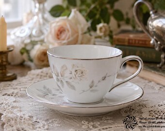 May include: A white porcelain teacup and saucer set featuring a delicate grey rose floral pattern and a thin silver rim. The teacup has a curved handle and sits centered on the matching saucer, both displaying a smooth, glossy finish.