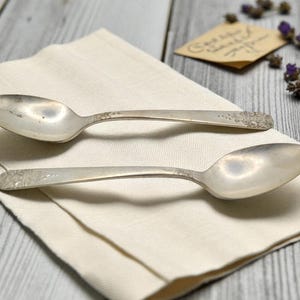 May include: Two vintage silver spoons with ornate handle detailing rest on a cream-colored linen napkin. The spoons are placed on a light gray wooden surface, with a small tag and lavender sprigs in the background.