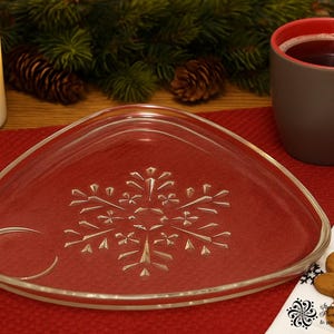 May include: A clear, triangular glass plate with a snowflake design, set on a red napkin. A brown mug with a red interior sits nearby, along with gingerbread cookies and a lit candle. Festive holiday theme.