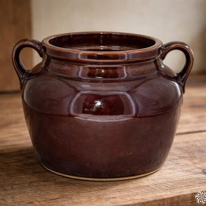 May include: A glossy, brown ceramic jar with two handles. The jar has a wide opening and a rounded body. It sits on a wooden surface. The jar is likely used for storage or decoration.