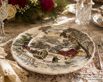 May include: A round ceramic dinner plate featuring a detailed winter landscape illustration. The scene depicts a snow-covered rural setting with a red barn, a horse-drawn carriage, and bare trees. The plate has a scalloped edge and a cream-colored base.