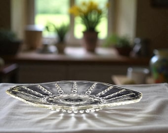 May include: A clear, decorative glass dish with a unique shape. The dish has a central circular design with radiating lines and beaded edges. It sits on a white cloth, with a blurred background of a kitchen and plants.