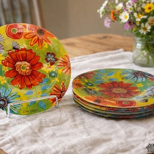 May include: A set of decorative plates with a floral design. The plates feature a vibrant mix of colors, including red, orange, blue, and yellow, with a green background. The plates are displayed on a white linen cloth.