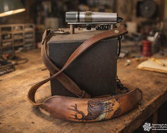 May include: A brown leather camera strap with a hand-tooled design of a creature and tree. The strap is draped over a dark box with a silver and black microphone attached. The items are on a wooden surface.