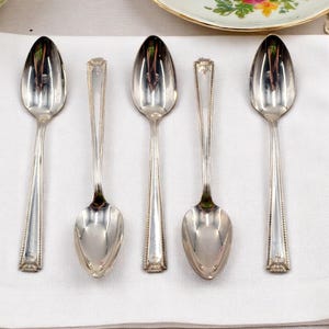 May include: Five silver spoons with oval bowls and ornate handles are arranged on a white cloth. The spoons have a reflective surface, and the handles feature a decorative pattern. The spoons are likely for serving or eating.