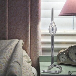May include: A clear glass decorative piece with a bulbous center and curved base stands on a mint green nightstand. A vintage clock and a lamp with a pink shade are also on the nightstand. The background includes a bed and a window.