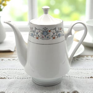 May include: A white porcelain teapot with a lid, featuring a floral design in blue, orange, and gray. The teapot has a curved spout and a handle. The background includes a window and other white teacups.